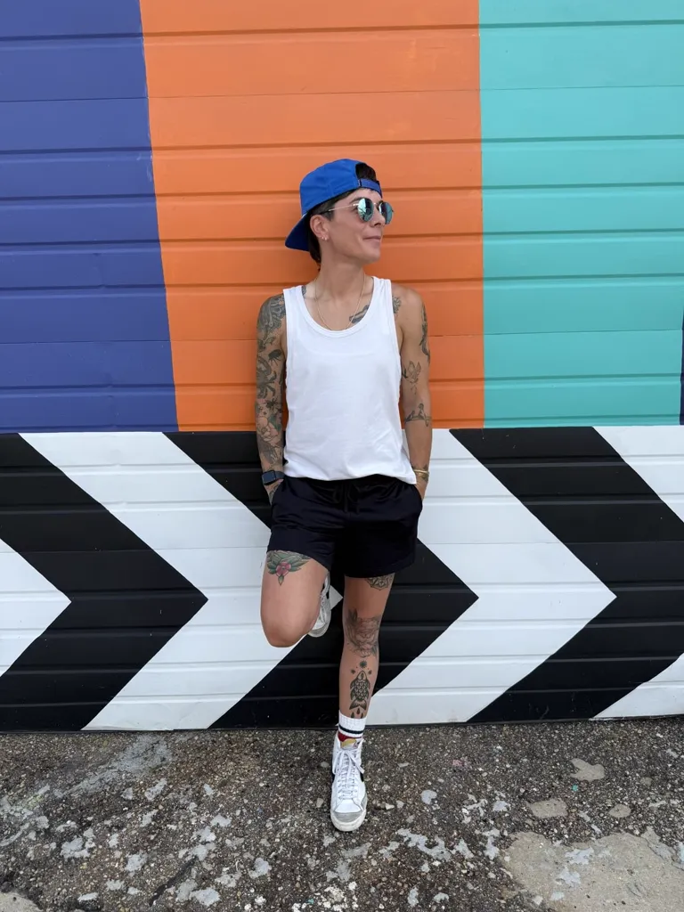 Kelly Schexnaildre in a backwards blue cap, round sunglasses, white tank top, and black shorts, leaning against a mural of purple, orange, and teal stripes above black-and-white chevrons.